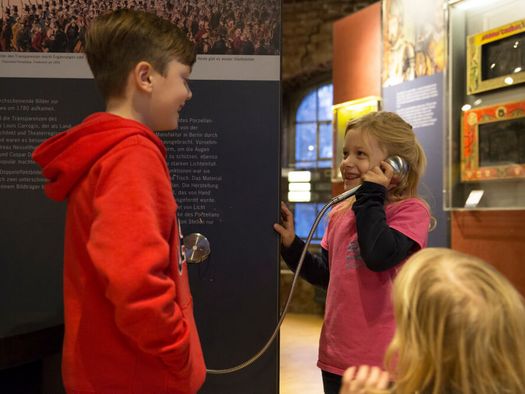 Kinder an einem Hörer mit Audioinfos im Museum zur Vorgeschichte des Films in der Camera Obscura Mülheim